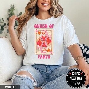 A woman wearing a white t-shirt with a graphic design of a woman holding a deck of playing cards and the text "Queen of Ears" printed on it.