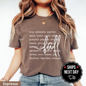 May include: A dark brown short-sleeve t-shirt featuring white text that lists fall-themed words including cozy sweaters, scarves, jeans, boots, apple cider, pumpkin patches, crunchy leaves, giving thanks, football, soup, harvest, candycorn, caramel apples, turkey, corn mazes, pie, bonfires, hayrides, and autumn, with the word fall written in a large white script font.