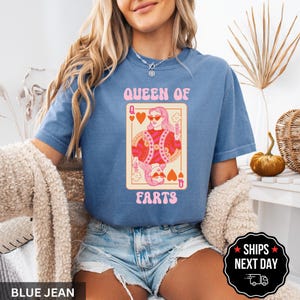 A woman wearing a blue t-shirt with a graphic design of a woman holding a sign that says "Queen of Farts" and denim shorts. She is sitting in a chair with a beige blanket draped over her.