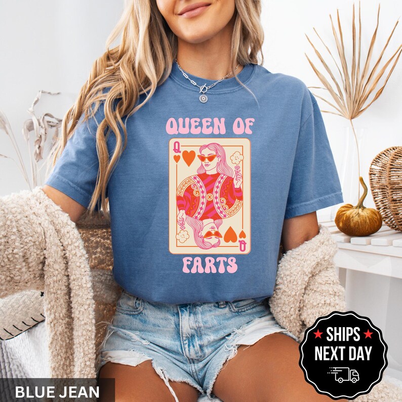 A woman wearing a blue t-shirt with a graphic design of a woman holding a sign that says "Queen of Farts" and denim shorts. She is sitting in a chair with a beige blanket draped over her.