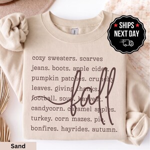 May include: A sand-colored crewneck sweatshirt featuring dark brown text listing fall-themed items such as cozy sweaters, scarves, jeans, boots, apple cider, pumpkin patches, crunchy leaves, giving thanks, football, soup, harvest, candycorn, caramel apples, turkey, corn mazes, pie, bonfires, hayrides, autumn, with the word fall written in large cursive script.