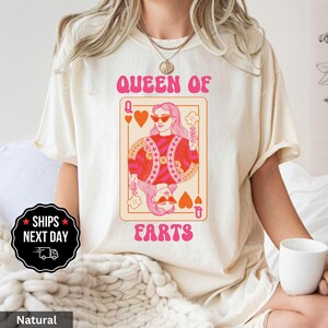 A woman wearing a white t-shirt with a graphic design of a woman's face and the text "Queen of Farts" printed on it.