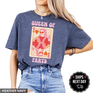 A woman wearing a blue t-shirt with a graphic design of a woman holding a deck of cards and the text "Queen of Farts" printed on it.