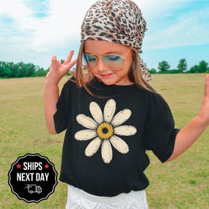 A young girl wearing a leopard print bandana, sunglasses, and a black t-shirt with a white flower design stands in a grassy field.