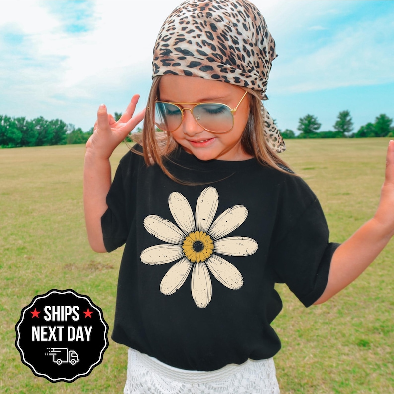 A young girl wearing a leopard print bandana, sunglasses, and a black t-shirt with a white flower design stands in a grassy field.