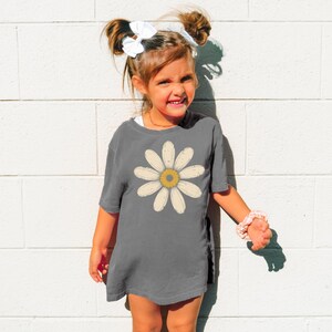 A young girl with a flower design on her shirt is smiling and posing against a white brick wall.