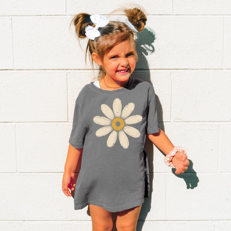 A young girl with a flower design on her shirt is smiling and posing against a white brick wall.