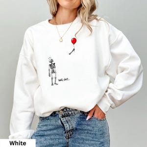 May include: A white long-sleeve sweatshirt featuring a graphic print of a skeleton looking up at a detached skeletal hand holding a red balloon. The text "Well, shit..." is printed in black ink next to the skeleton figure on the front of the garment.