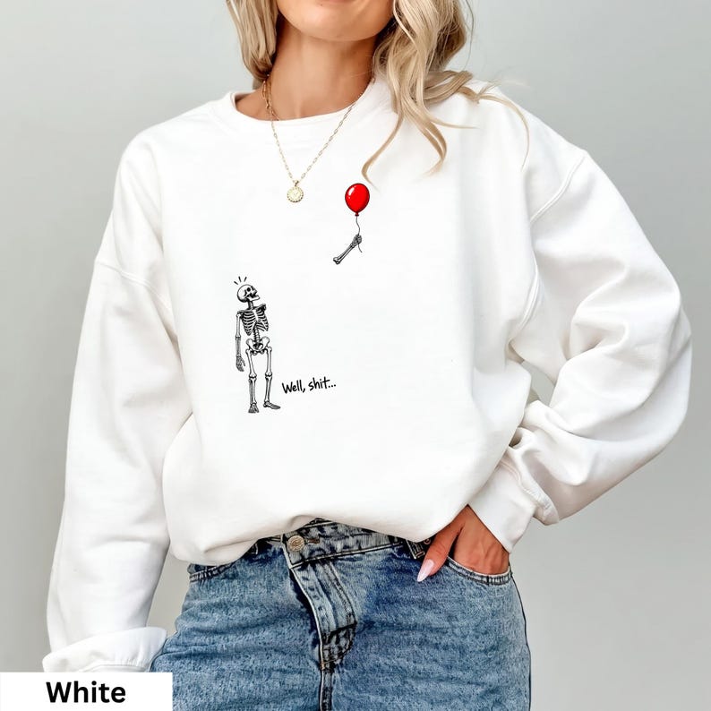 May include: A white long-sleeve sweatshirt featuring a graphic print of a skeleton looking up at a detached skeletal hand holding a red balloon. The text "Well, shit..." is printed in black ink next to the skeleton figure on the front of the garment.