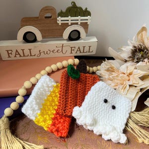 May include: A collection of handmade crochet fall-themed decorations. Includes a candy corn, a pumpkin, and a ghost. A wooden truck with pumpkins and the text "FALL sweet FALL" are in the background. The decorations are in shades of white, orange, yellow, and green.
