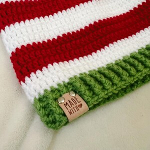 Pu&ograve; includere: Un cappello all'uncinetto fatto a mano con strisce rosse e bianche alternate. Il bordo &egrave; verde e una piccola etichetta in pelle con le parole "MADE WITH" e un cuore &egrave; attaccata. Il cappello &egrave; probabilmente per l'inverno.