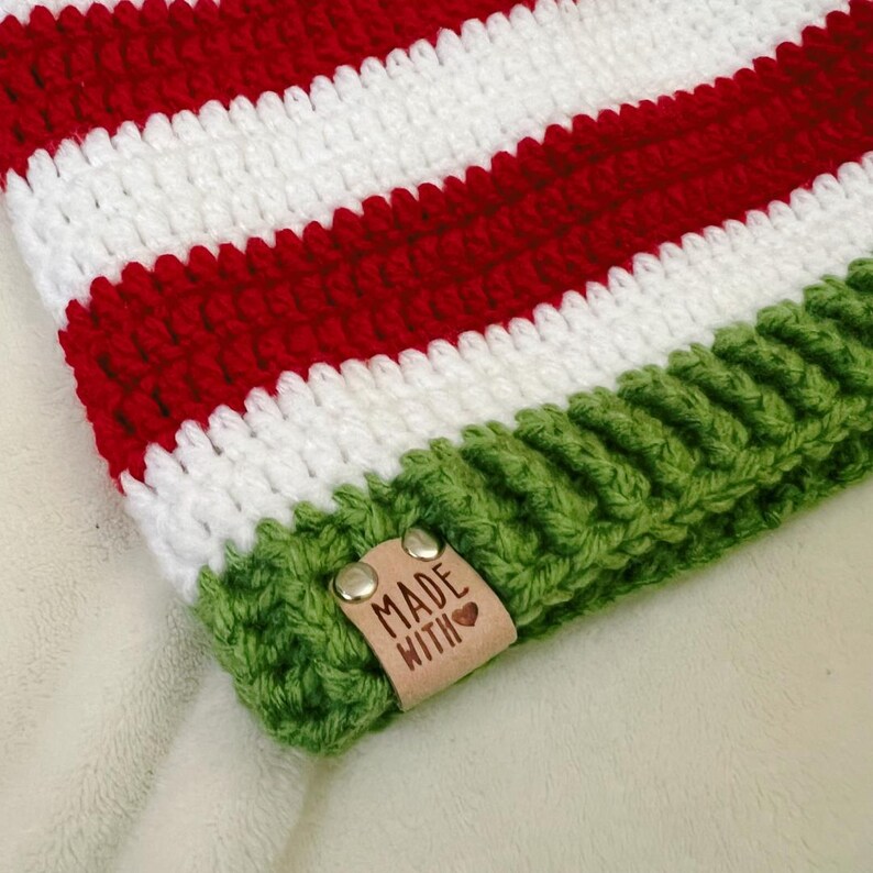Pu&ograve; includere: Un cappello all'uncinetto fatto a mano con strisce rosse e bianche alternate. Il bordo &egrave; verde e una piccola etichetta in pelle con le parole "MADE WITH" e un cuore &egrave; attaccata. Il cappello &egrave; probabilmente per l'inverno.
