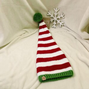 Pu&ograve; includere: Un cappello da elfo fatto a mano, a righe rosse e bianche, con un pompon verde e un bordo verde. Il cappello &egrave; lavorato all'uncinetto e ha una piccola etichetta con la scritta "Made With". Il cappello &egrave; esposto su una superficie bianca strutturata.