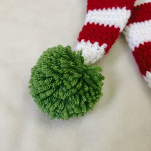 Pu&ograve; includere: Primo piano di un pompon verde e di un design all'uncinetto a righe rosse e bianche. Il motivo all'uncinetto &egrave; un design festivo per le vacanze. Il pompon &egrave; di colore verde brillante ed &egrave; fatto di filato morbido.