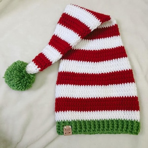 Pu&ograve; includere: Un cappello da elfo fatto a mano all'uncinetto con strisce rosse e bianche alternate. Il cappello presenta un pompon verde sulla punta e un bordo verde. Una piccola etichetta con la scritta "MADE WITH" &egrave; attaccata al bordo.