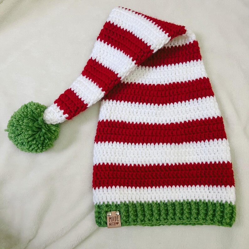 Pu&ograve; includere: Un cappello da elfo fatto a mano all'uncinetto con strisce rosse e bianche alternate. Il cappello presenta un pompon verde sulla punta e un bordo verde. Una piccola etichetta con la scritta "MADE WITH" &egrave; attaccata al bordo.