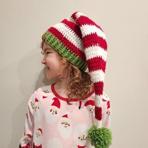 Pu&ograve; includere: Un cappello da elfo a righe rosse e bianche con bordo verde e pompon verde. Il cappello &egrave; all'uncinetto e ha una forma lunga e appuntita. La persona indossa un top pigiama rosa con un motivo di Babbo Natale.