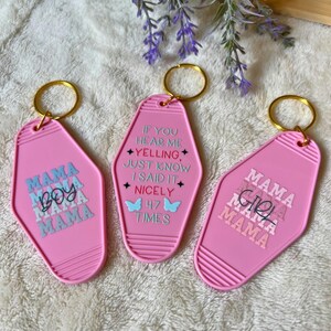 May include: Three pink keychains with gold key rings. One keychain says "MAMA BOY MAMA MAMA" in blue and gray text. Another says "IF YOU HEAR ME YELLING JUST KNOW I SAID IT NICELY 47 TIMES". The third says "MAMA GIRL MAMA MAMA".
