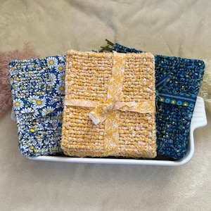 May include: Three crocheted pot holders in a white ceramic dish. One pot holder is yellow with a white floral pattern, one is blue with a white daisy pattern, and one is blue with a white geometric pattern.