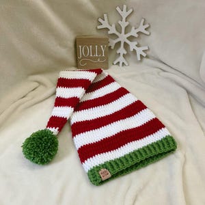 Pu&ograve; includere: Un cappello da elfo all'uncinetto fatto a mano con strisce rosse e bianche, un pompon verde e un bordo verde. Un blocchetto di legno con la scritta "JOLLY" &egrave; sullo sfondo, insieme a un fiocco di neve bianco.