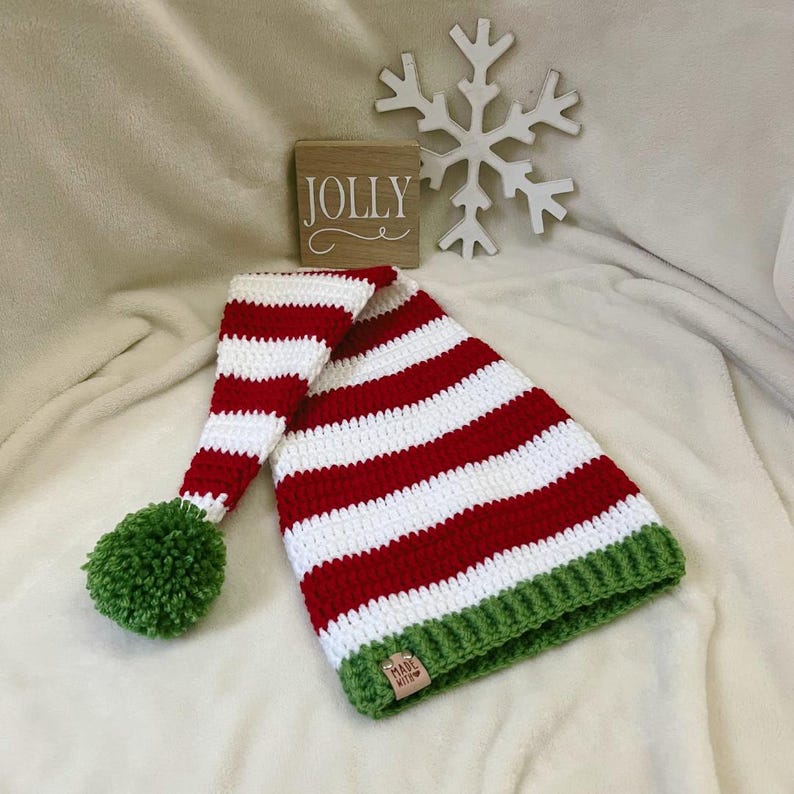 Pu&ograve; includere: Un cappello da elfo all'uncinetto fatto a mano con strisce rosse e bianche, un pompon verde e un bordo verde. Un blocchetto di legno con la scritta "JOLLY" &egrave; sullo sfondo, insieme a un fiocco di neve bianco.