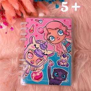 Può includere: Un quaderno a spirale in plastica trasparente con un design di cartoni animati colorati che presenta una ragazza con i capelli rosa, una ragazza vestita con un costume da unicorno, un unicorno e un gatto nero. Il quaderno ha il testo "5+" sulla copertina.