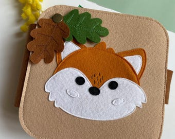 May include: A tan felt book cover with a fox face design in orange, white, and black. The cover is adorned with brown and green felt leaves and a brass button. Yellow flowers and a light green surface are in the background.