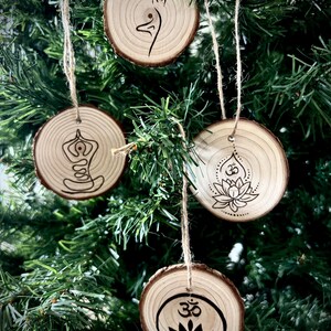 Könnte beinhalten: Vier Holzornamente mit einer natürlichen Holzoptik und schwarzen Designs. Die Ornamente zeigen Yoga-Posen und das Om-Symbol. Die Ornamente sind mit einer Schnur befestigt.