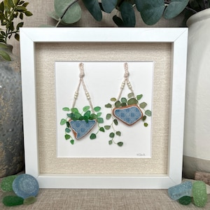 Puede incluir: Una obra de arte enmarcada con dos macetas colgantes hechas de vidrio de mar. Las macetas están decoradas con un patrón de azulejos azules y blancos y están llenas de vidrio de mar verde que representa plantas. La obra está firmada "K.Clark".