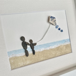 May include: Framed pebble art depicting a parent and child on a beach, flying a kite. The kite is white and blue, with a string of blue beads. The beach is tan with blue watercolor waves. The frame is white.