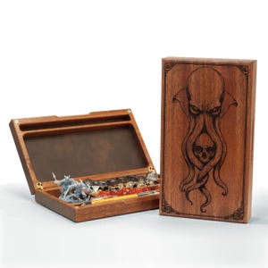 May include: A wooden dice box with an open lid, revealing dice and miniature figures. The box is next to a wooden lid with an engraved image of an octopus-like creature and a skull. The box and lid are made of a dark wood.