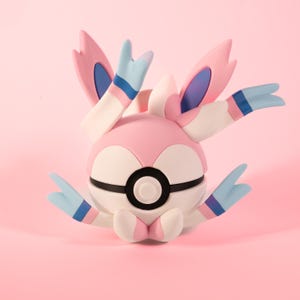 Könnte beinhalten: Eine rosafarbene und weiße Pokémon-Figur eines Pokéballs mit einem blauen und weißen Schwanz und Ohren. Die Figur sitzt auf einem rosa Hintergrund.