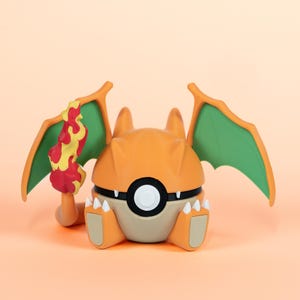 Könnte beinhalten: Orange-braune Charizard-Pokéball-Figur mit grünen Flügeln und einem Flammen-Accessoire. Die Figur hat ein schwarz-weißes kreisförmiges Design auf der Vorderseite und ist vor einem pfirsichfarbenen Hintergrund platziert.
