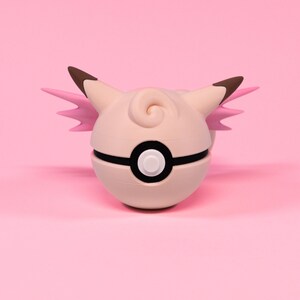 Könnte beinhalten: Ein hellrosa, kugelförmiges Objekt mit einem schwarzen Streifen und einem weißen Knopf, das einem Pokéball ähnelt. Es zeigt einen hellrosa, cartoonartigen Charakter mit braunspitzigen Ohren und rosa flügelartigen Details. Der Hintergrund ist einfarbig hellrosa.