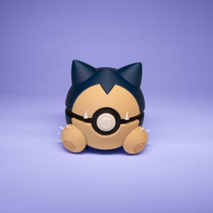 Könnte beinhalten: Eine blaue und braune Pokémon-Figur eines runden, zusammengerollten Wesens mit einem weißen Kreis auf der Brust. Die Figur hat spitze Ohren und kleine, braune Pfoten.