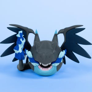 Könnte beinhalten: Stilisierte Figur einer drachenartigen Kreatur mit grauem Kopf, dunkelblauen Flügeln und blau-weißen Akzenten. Die Kreatur hat ein offenes Maul mit sichtbaren Zähnen, vor einem hellblauen Hintergrund.