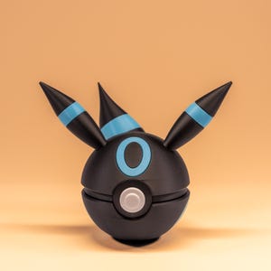 Könnte beinhalten: Eine schwarz-blaue Umbreon-Pokéball-Replik. Das kugelförmige Design hat einen schwarzen Körper mit blauem Ring und Akzenten sowie drei spitze Ohren mit blauen Streifen. Ein weißer Knopf befindet sich mittig auf der Vorderseite.