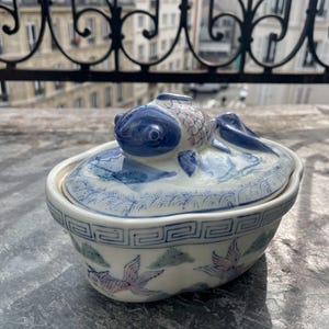 Puede incluir: Un plato de cerámica para servir con tapa, con un diseño de pez azul y blanco. El plato tiene forma rectangular con bordes redondeados y está decorado con motivos de peces y nubes. La tapa está rematada con una figura de pez azul.