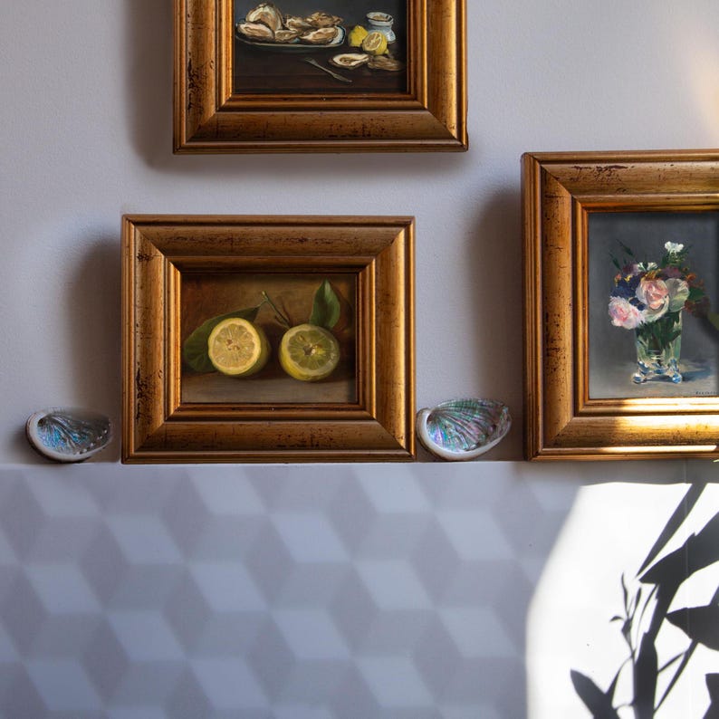 Op de afbeelding: Drie ingelijste schilderijen met goudkleurige lijsten hangen aan een lichtgrijze muur. De schilderijen tonen stillevens, waaronder citroenen, oesters en bloemen in een vaas. Twee iriserende schelpvormige schalen zijn eronder geplaatst.