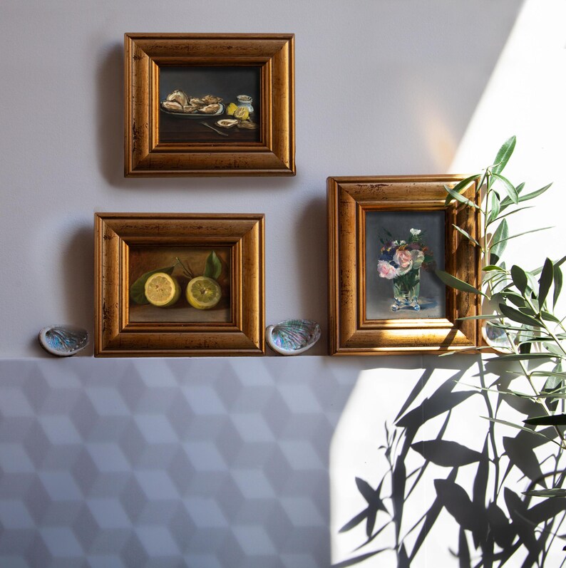 Op de afbeelding: Drie ingelijste olieverfschilderijen met goudkleurige lijsten worden aan een muur getoond. De schilderijen tonen een stilleven van citroenen, een vaas met bloemen en een bord met eten. Twee decoratieve schelpen zijn eronder geplaatst.
