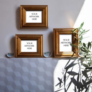 Op de afbeelding: Drie vierkante, goudkleurige fotolijsten met de tekst "YOUR ARTWORK HERE" worden weergegeven op een lichtgrijze muur. De lijsten zijn in een cluster gerangschikt, met twee lijsten onder &eacute;&eacute;n. Decoratieve schelpen zijn onder de lijsten geplaatst.