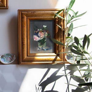 Op de afbeelding: Een ingelijst bloemstilleven schilderij. Het kunstwerk toont een boeket van roze en witte bloemen in een heldere glazen vaas. De lijst is goudkleurig. Het schilderij wordt op een witte muur getoond.