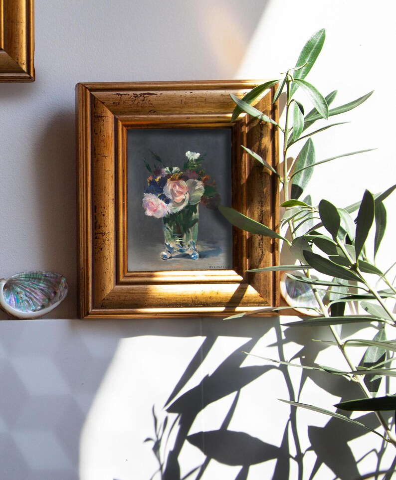 Op de afbeelding: Een ingelijst bloemstilleven schilderij. Het kunstwerk toont een boeket van roze en witte bloemen in een heldere glazen vaas. De lijst is goudkleurig. Het schilderij wordt op een witte muur getoond.