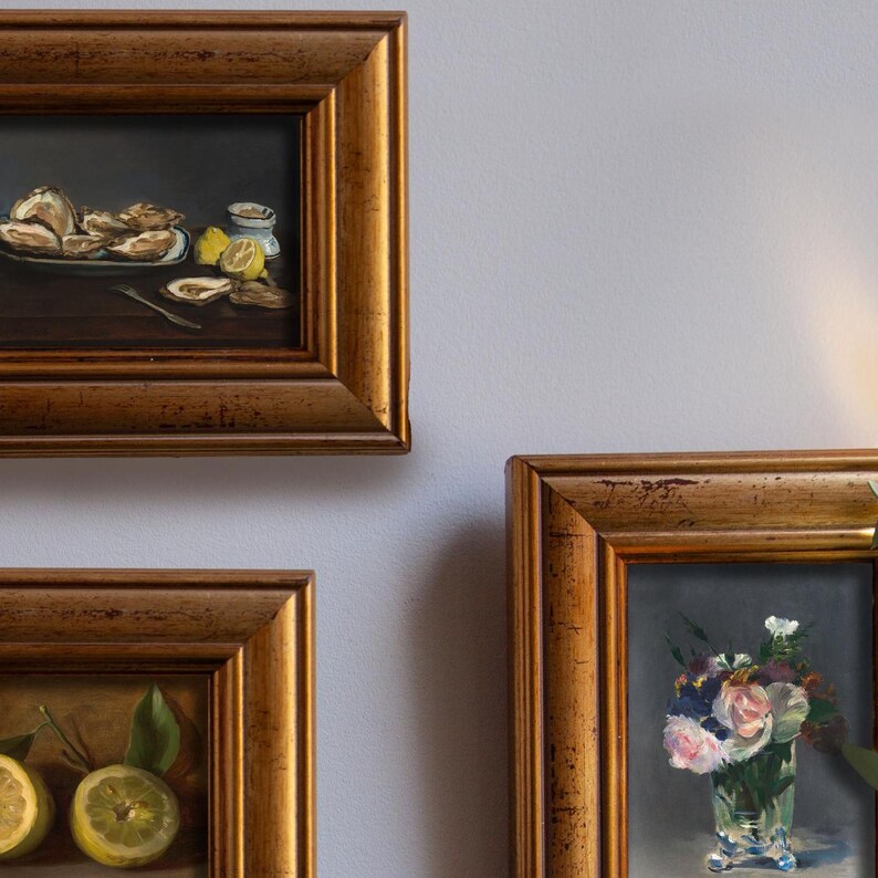 Op de afbeelding: Drie ingelijste stillevens met goudkleurige lijsten. De schilderijen tonen oesters op een bord met citroenen, citroenen met bladeren en een vaas met bloemen. De schilderijen worden tentoongesteld tegen een lichtgrijze muur.
