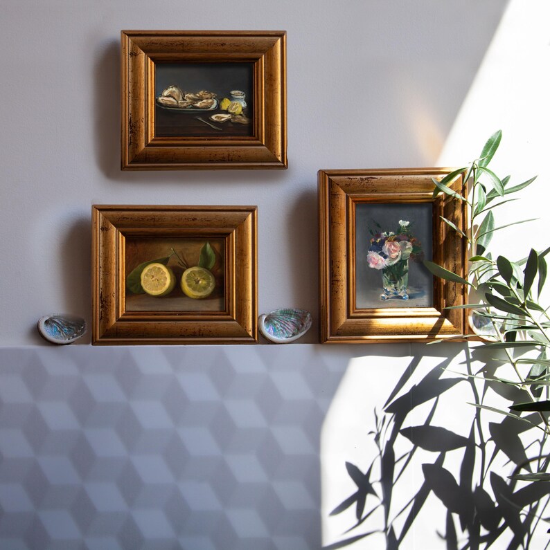 Op de afbeelding: Drie ingelijste olieverfschilderijen met goudkleurige lijsten worden aan een muur getoond. De schilderijen tonen stillevens van oesters, citroenen en bloemen in een vaas. Twee decoratieve schelpen zijn onder de schilderijen geplaatst en een plant staat in de hoek.