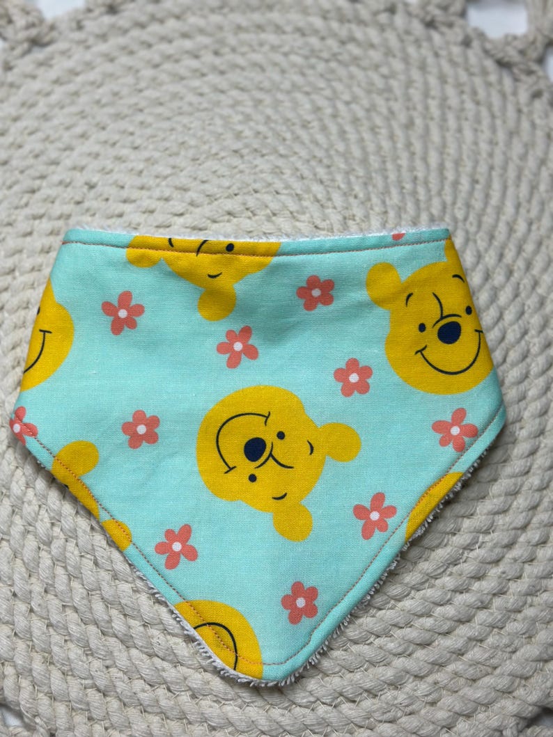 May include: A light blue baby bib with a Winnie the Pooh design. The bib features a repeating pattern of Winnie the Pooh's face and pink flowers.