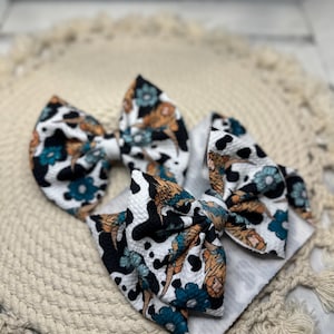 May include: Three fabric hair bows with a cow print design, featuring black spots, teal and orange floral accents, and a white background. The bows are arranged on a cream-colored, textured surface.