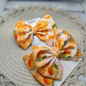 May include: Two orange and white patterned hair bows with a citrus fruit design. The bows are made of fabric and have a velvet center. The bows are on a white card and are set on a beige, textured surface.