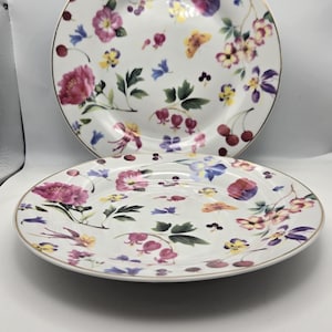 May include: Two white ceramic plates with a gold rim. The plates feature a floral pattern with pink, purple, yellow, and blue flowers on a white background.