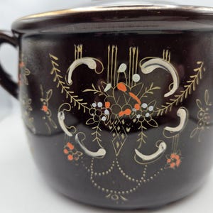 May include: A dark brown ceramic pot with two handles and a lid. The pot is decorated with a floral design in white, gold, and orange. The rim of the pot has a gold trim.